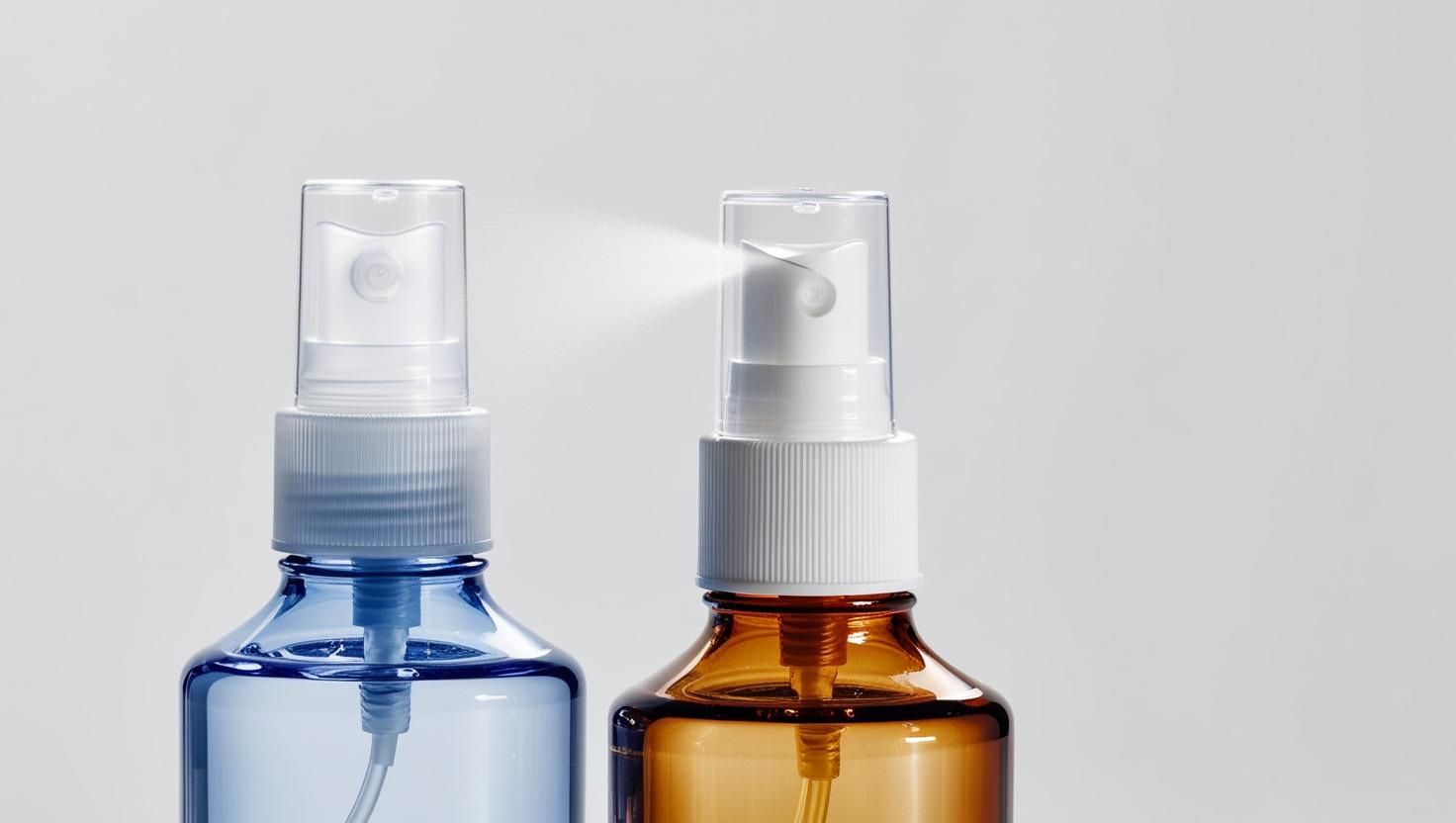 Close-up of blue and amber spray bottles, one emitting a fine mist, against a light background.